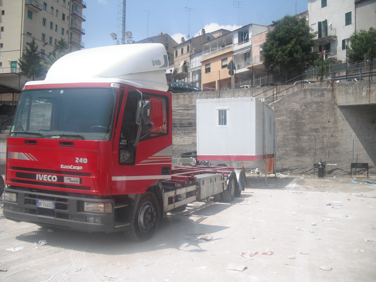 Camion rosso con il braccio che solleva e posa un container cucina bianco in un piazzale