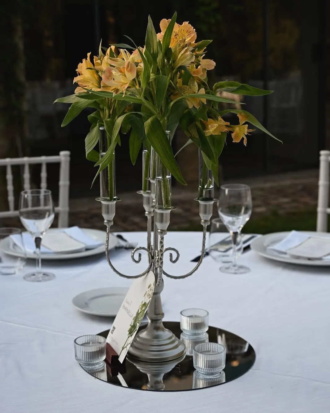 Centrotavola con candelabro argento, fiori gialli e portacandele in vetro su tavolo rotondo Buratti Catering