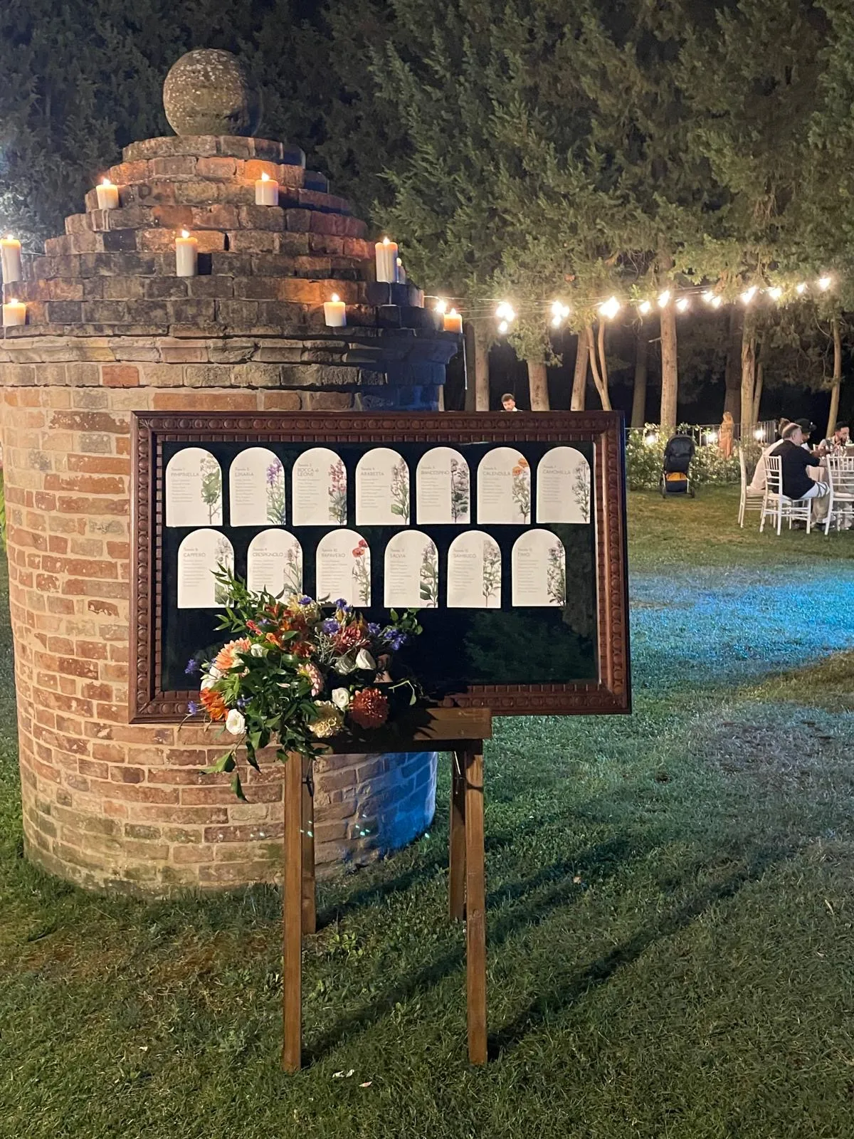 Tableau de mariage illuminato di sera con candele su struttura in mattoni e cartoncini dei tavoli decorati a tema botanico