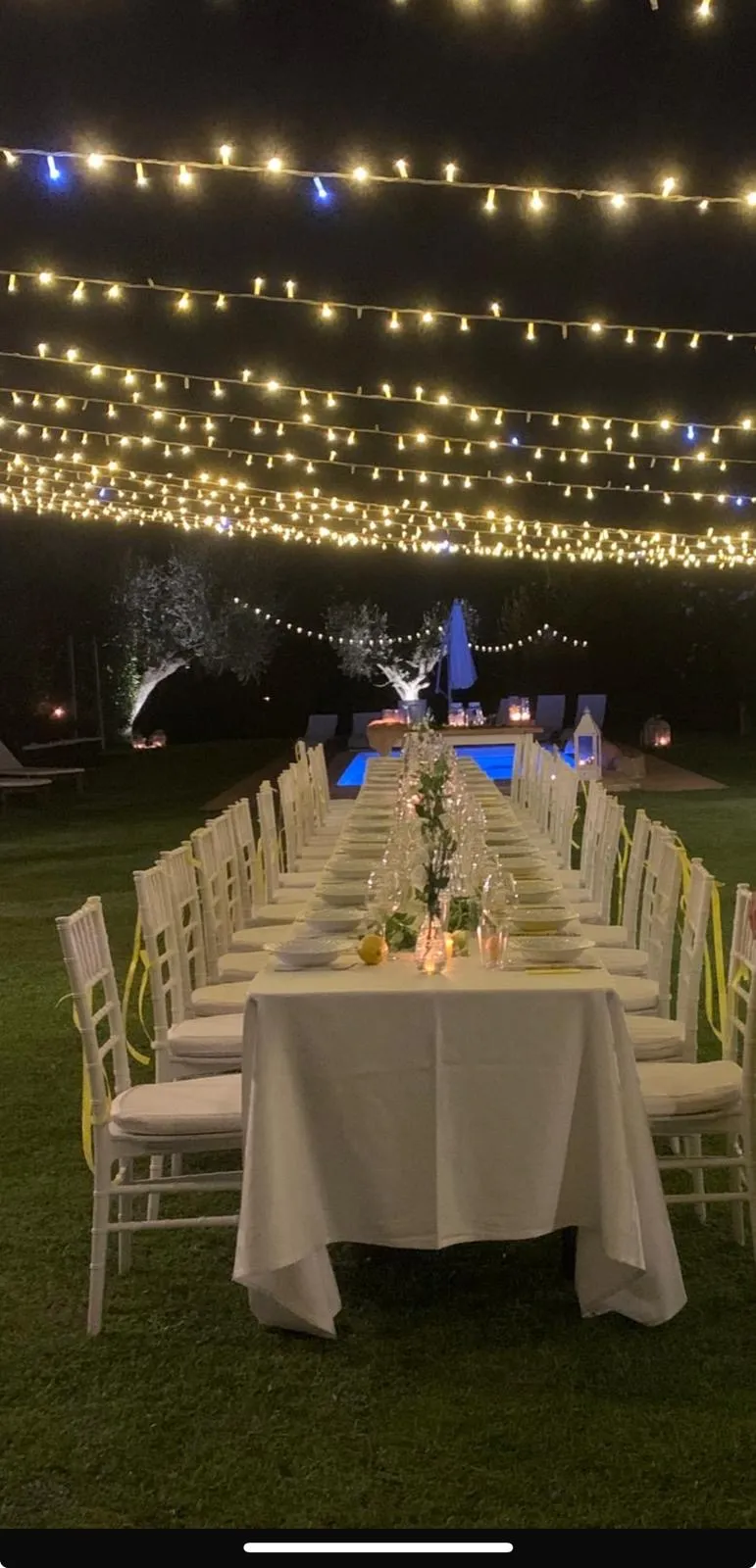 Lunga tavolata di sera con sedie bianche e fili di luci sopra per catering matrimonio all'aperto