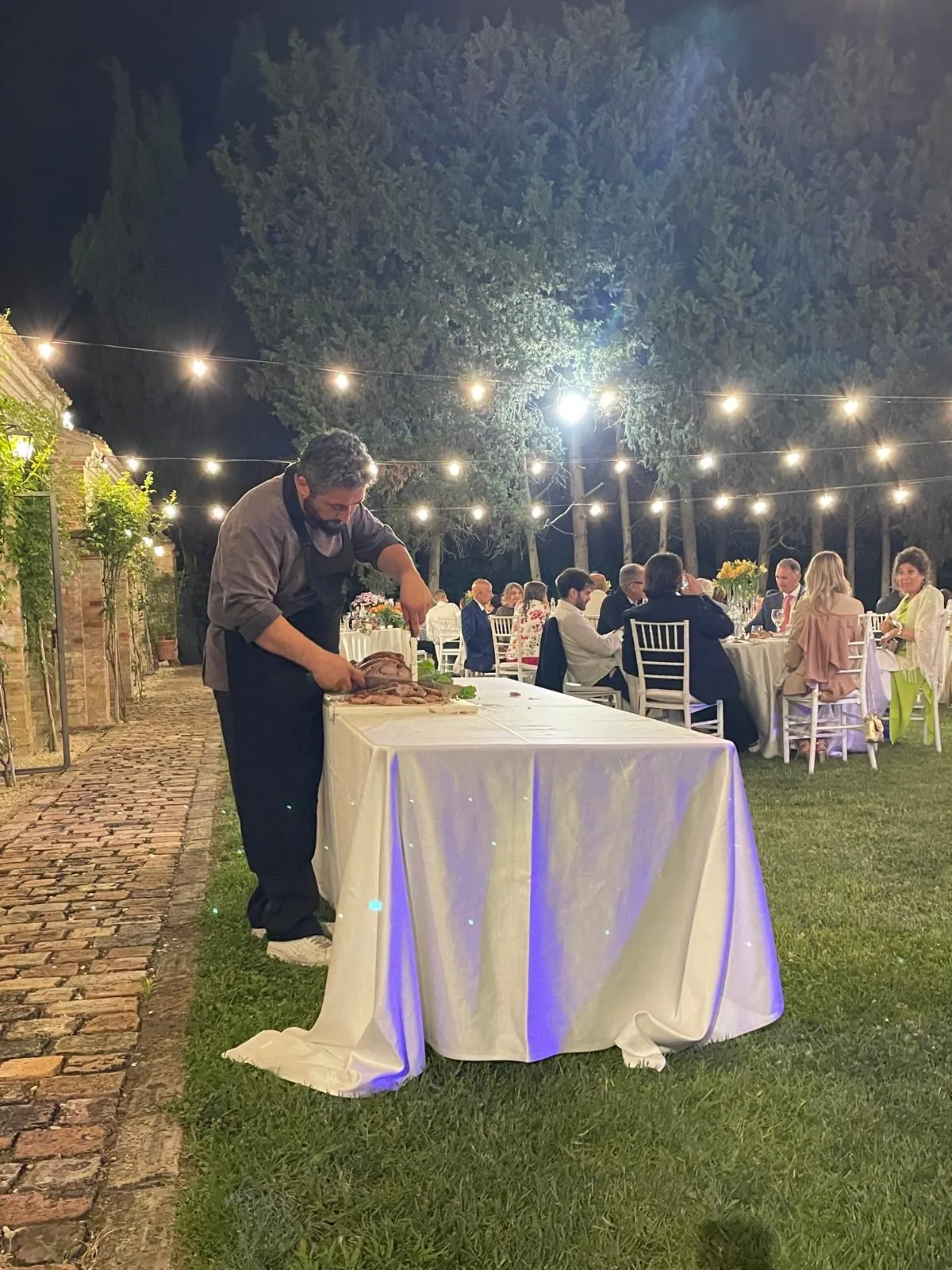 Chef che affetta carne su tavolo di servizio all'aperto di sera con luci sospese e ospiti seduti ai tavoli