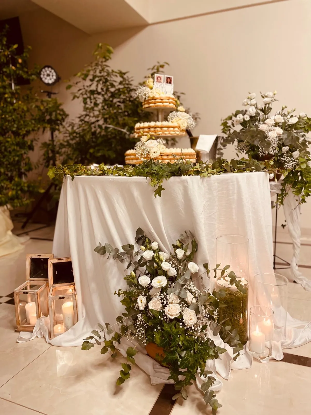 Tavolo torta nuziale a piani con fiori bianchi, candele e lanterne su tovaglia bianca