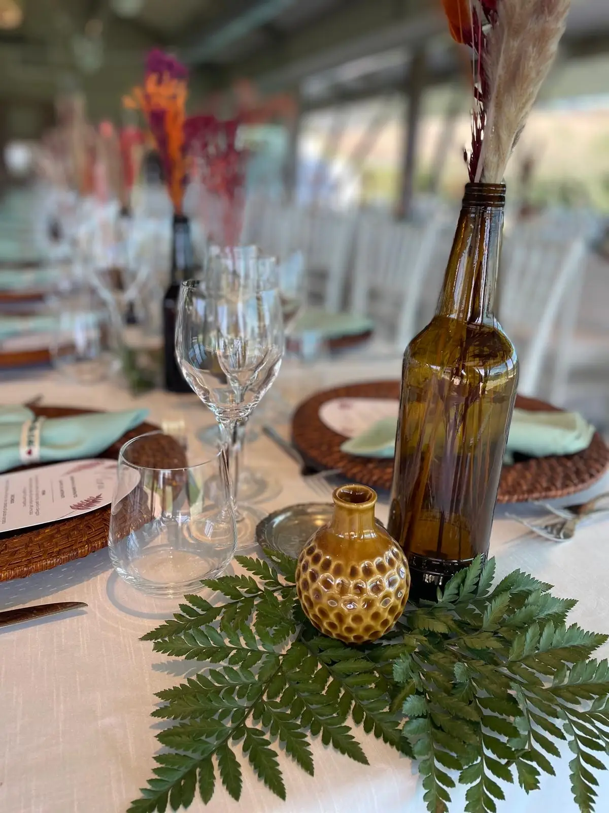 Dettaglio centrotavola con bottiglia ambrata, piccolo vaso senape e foglia di felce