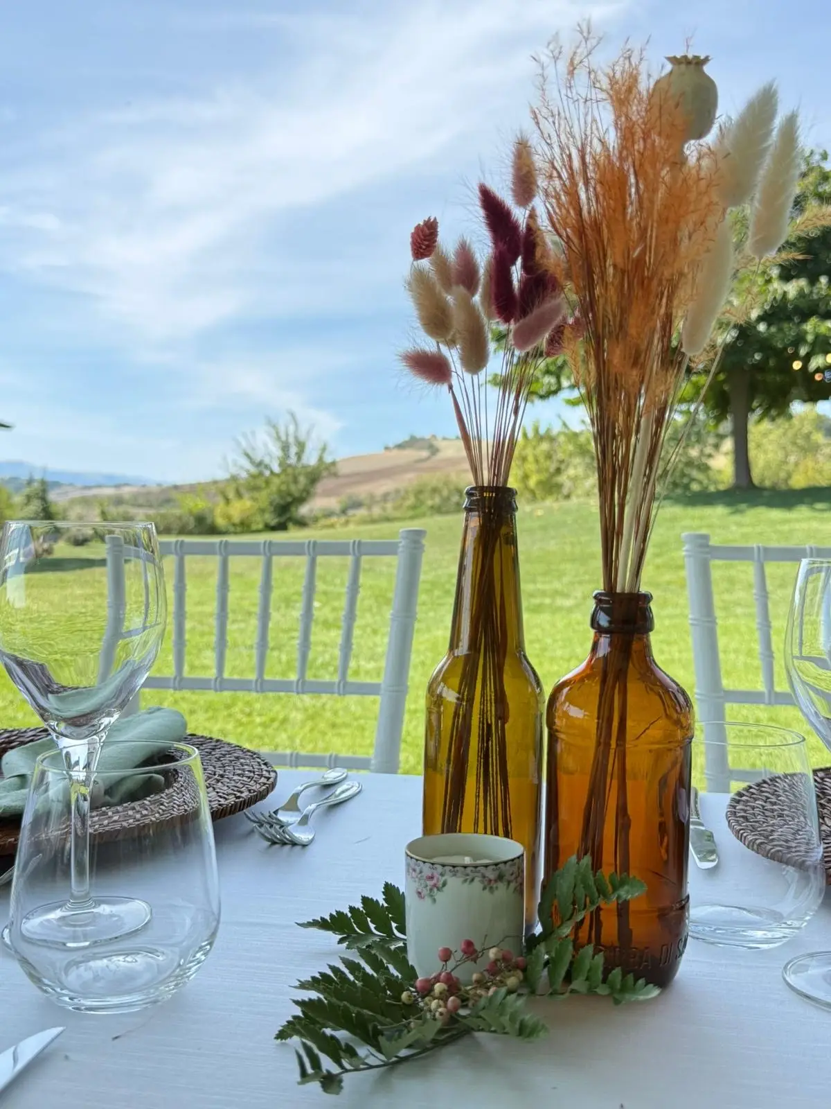 Dettaglio centrotavola con bottiglie ambrate, fiori secchi e foglia di felce