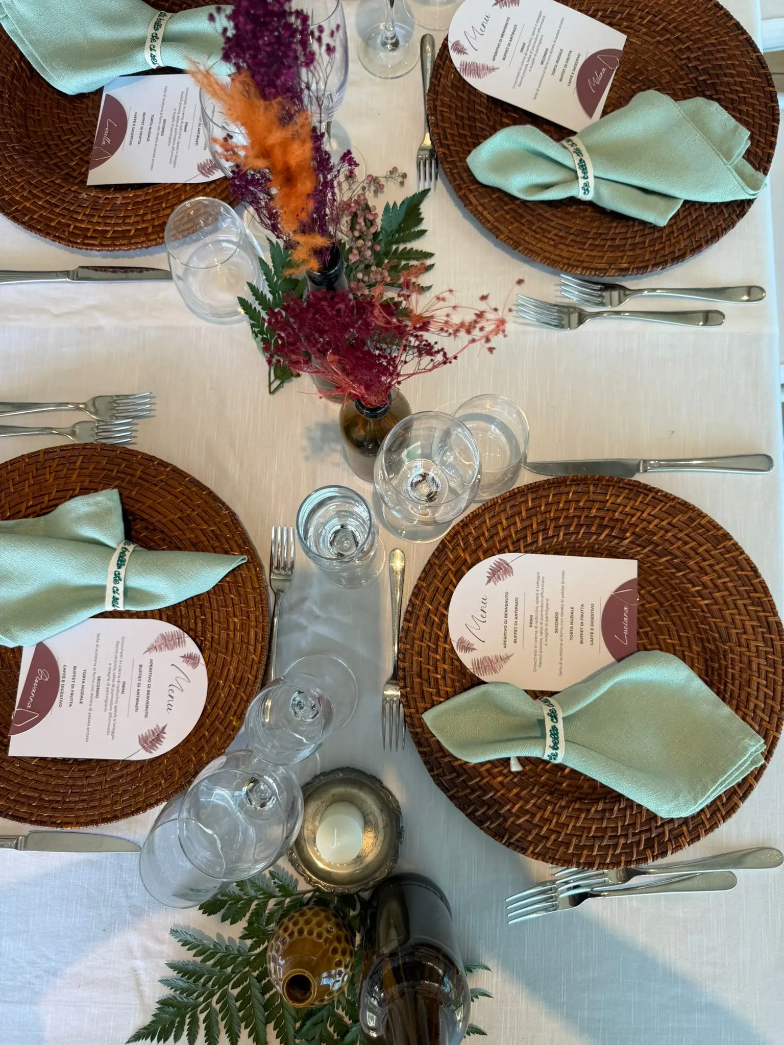 Mise en place vista dall'alto con piatti in rattan, tovaglioli verde salvia e menu coordinati