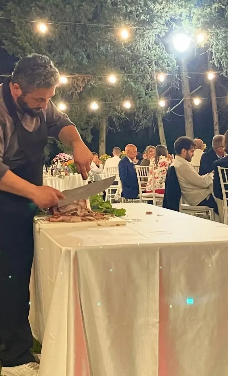Un cuoco taglia la carne su un tavolo con tovaglia chiara, di sera, sotto fili di lampadine, con gli ospiti seduti ai tavoli sullo sfondo