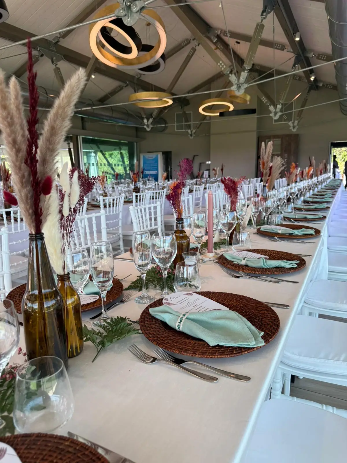 Lunga tavolata interna con sedie bianche, piatti in rattan e centrotavola in bottiglia