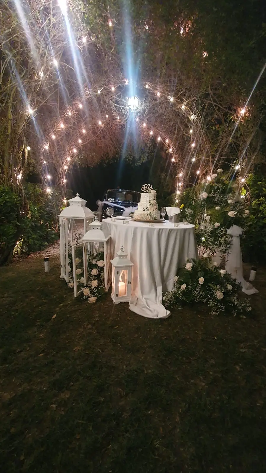 Tavolo della torta illuminato da archi di luci e circondato da lanterne bianche