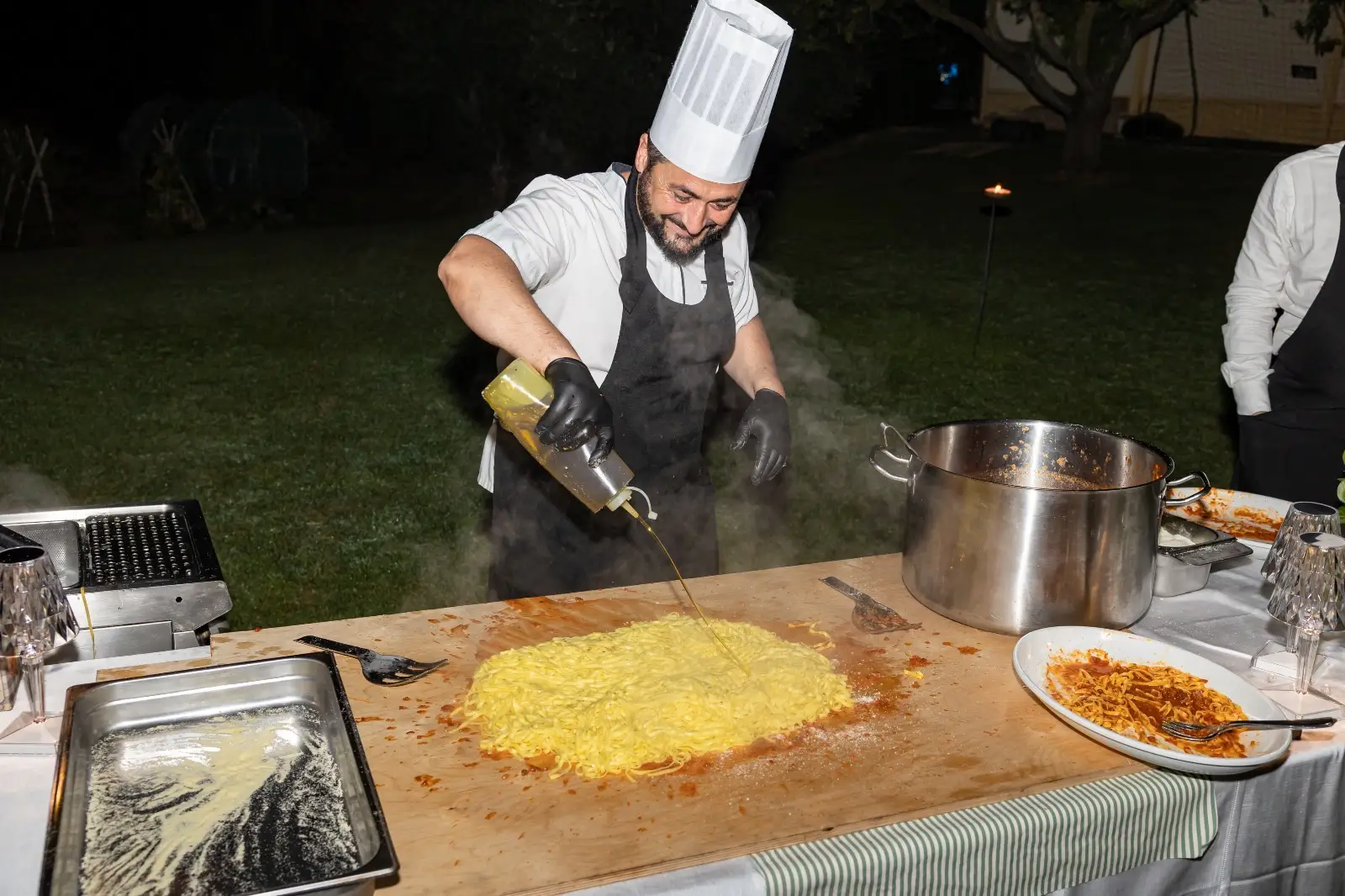 Chef che condisce pasta fumante su grande tavolo da lavoro all'aperto