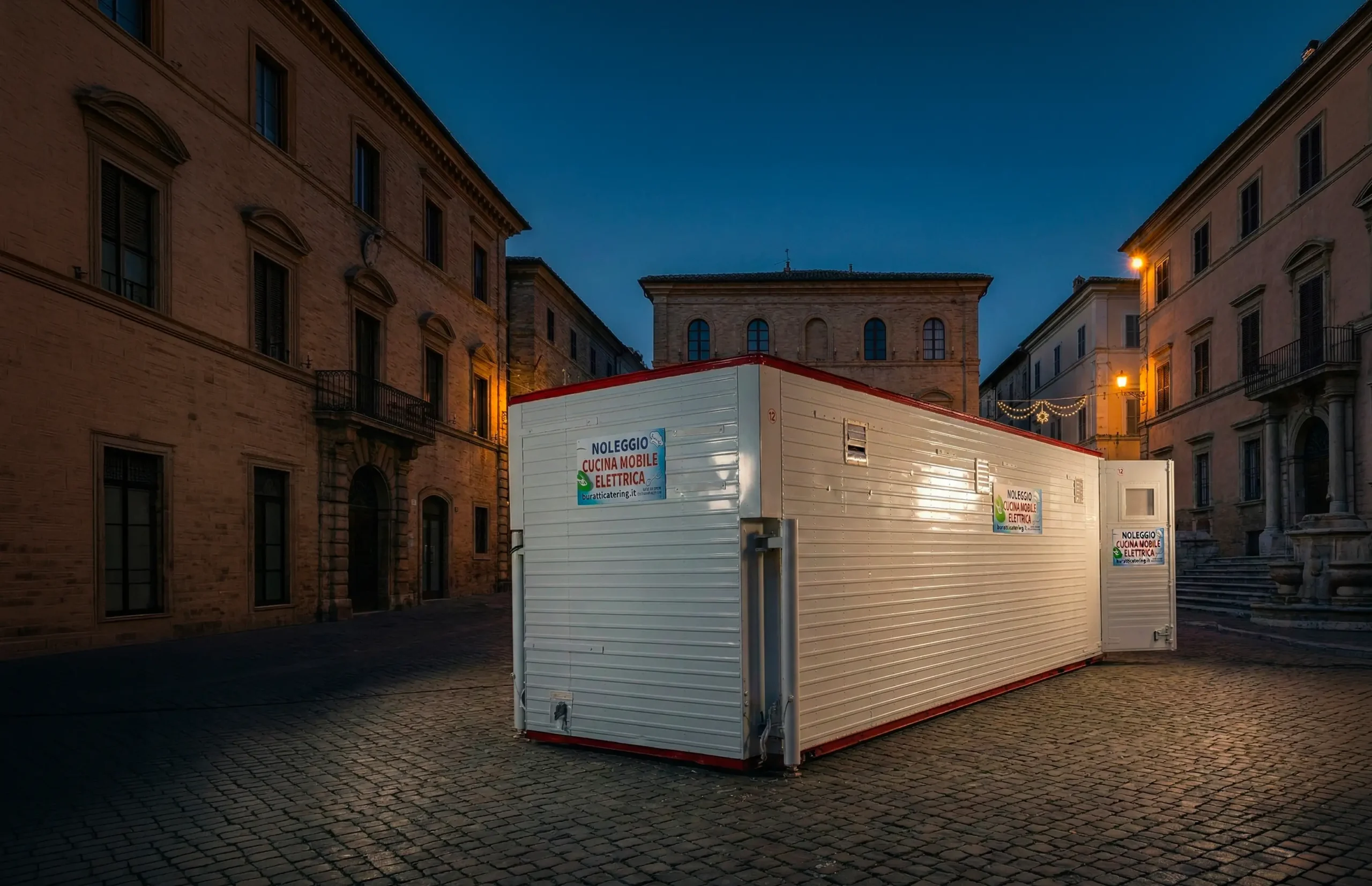 Cucina mobile container elettrica E-GO N.12 Buratti Catering illuminata di notte davanti a un borgo storico — noleggio cucine mobili professionali