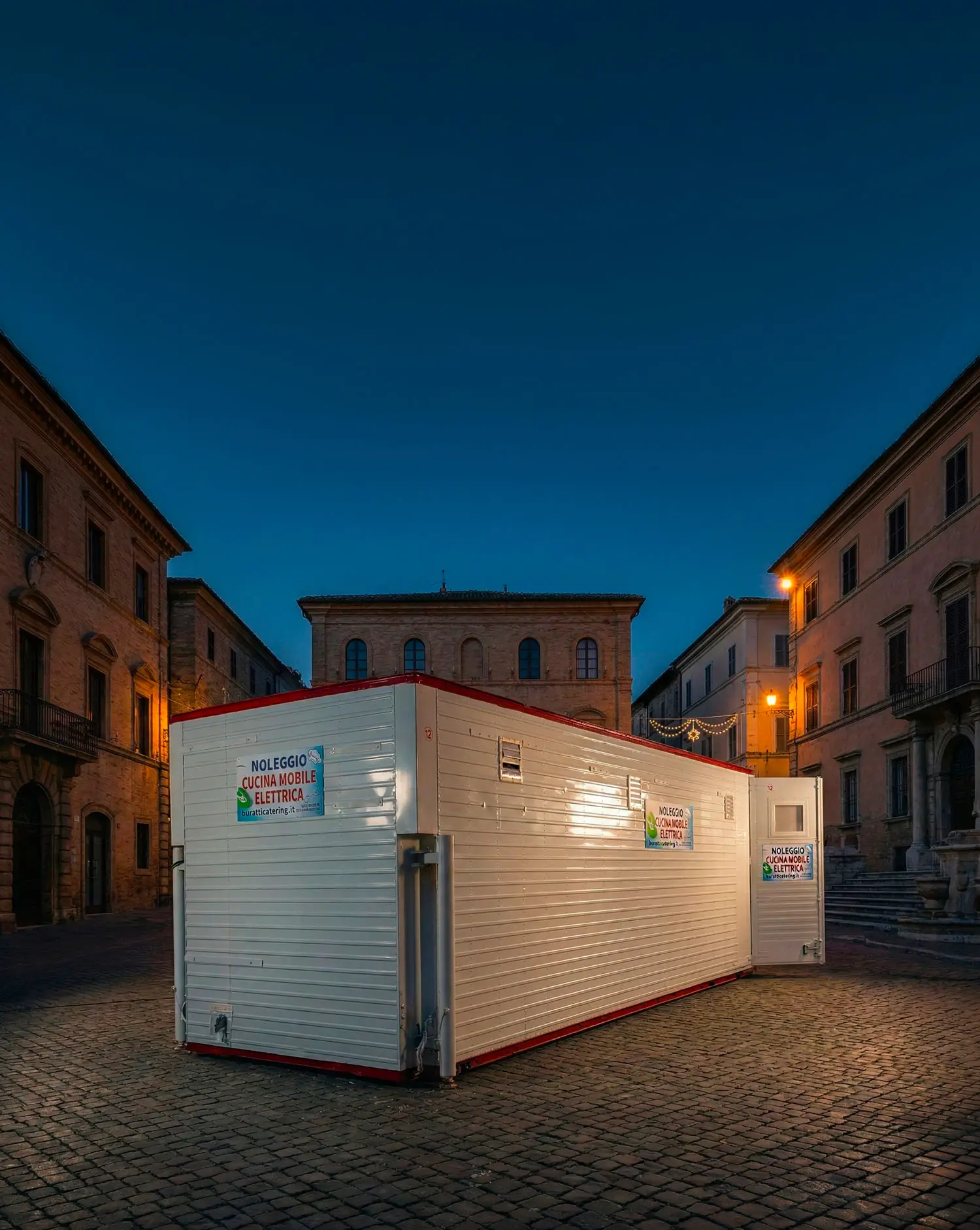 Cucina mobile elettrica E-GO 12 a noleggio per concerti e festival, illuminata di notte in un borgo storico italiano